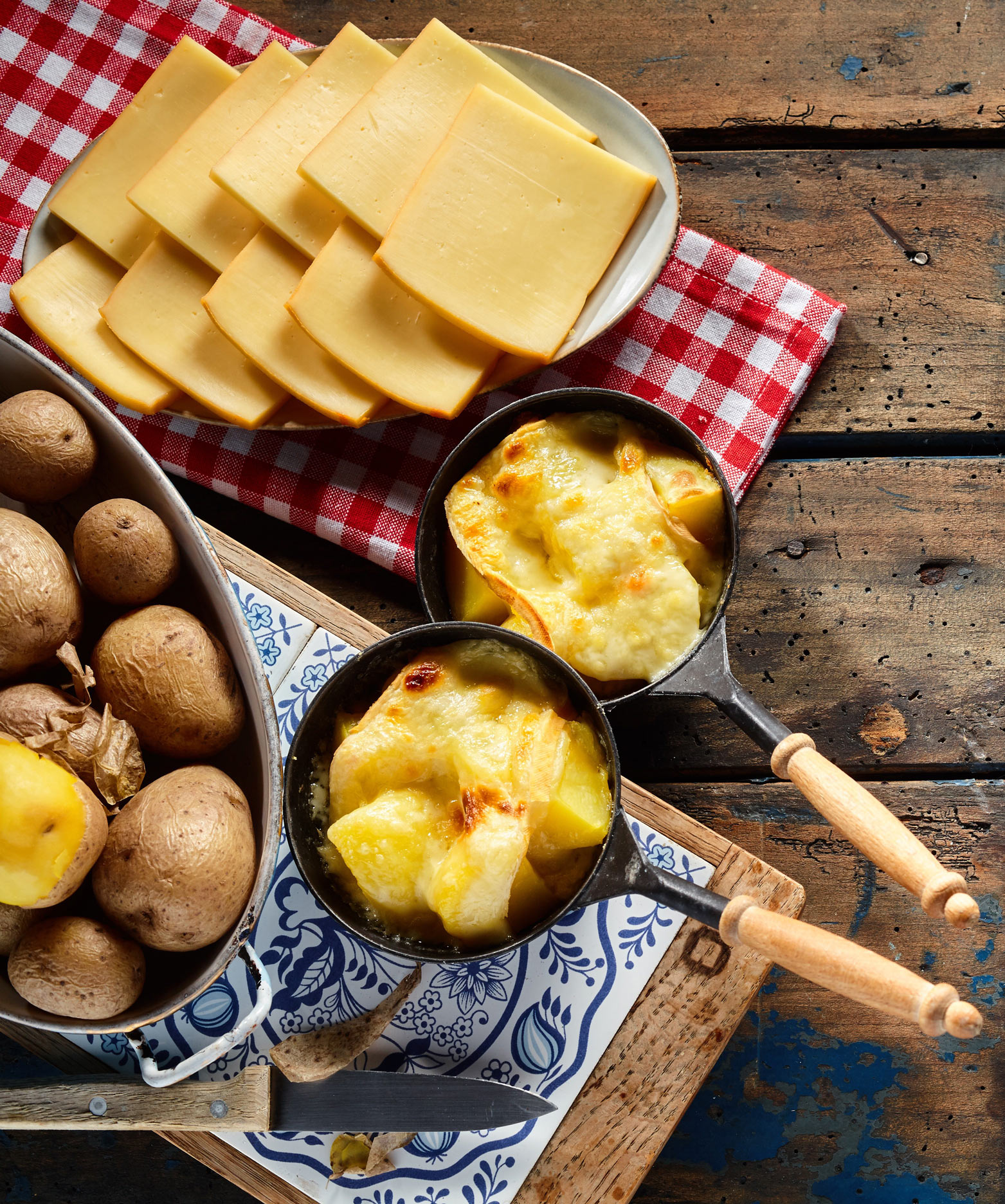 Raclettegericht mit geschmolzenem Käse in kleinen Pfännchen, serviert mit gekochten Kartoffeln und Käsescheiben auf kariertem Tuch, rustikal angerichtet auf Holztisch.