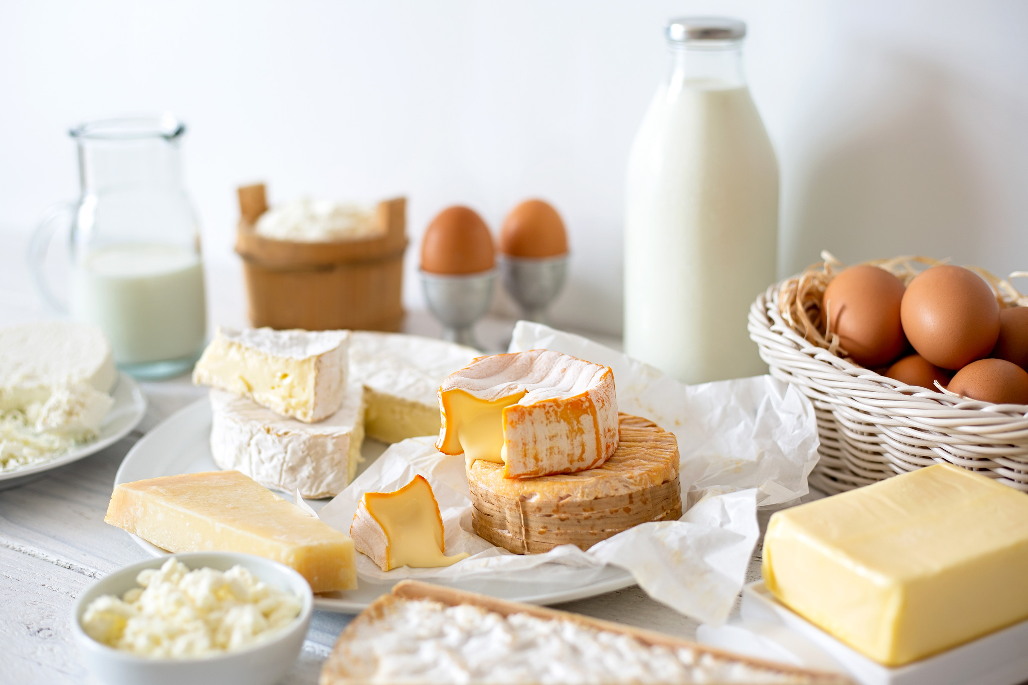 Auswahl an Käse, Milch und Eiern Verschiedene Käsesorten, Butter, Milch und Eier auf einem weissen Tisch angerichtet. Im Vordergrund liegen Weichkäse, Hartkäse und Frischkäse, im Hintergrund ein Korb mit braunen Eiern und eine Glasflasche Milch.