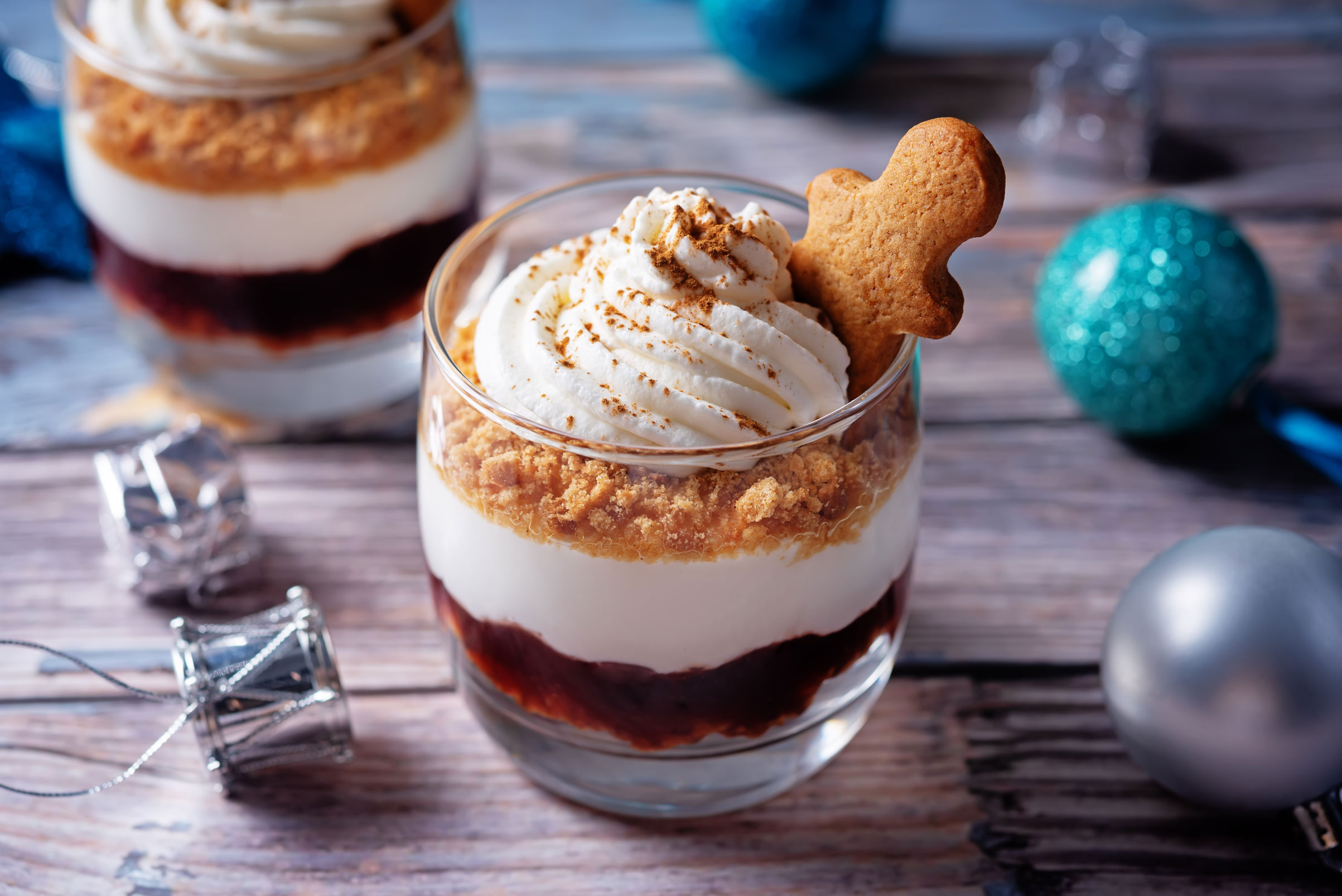 Ein winterliches Dessert im Glas mit geschichteter Milchcreme, Keksstreuseln, fruchtiger Sauce und einem kleinen Lebkuchen-Plätzchen, dekoriert mit Zimt. Im Hintergrund liegen weihnachtliche Kugeln und silberne Deko.