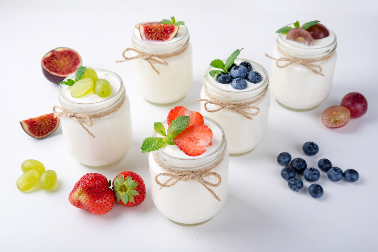 Mehrere Gläser mit cremigem Joghurt, dekoriert mit frischen Erdbeeren, Blaubeeren, grünen Weintrauben, Feigen und Minzblättern, rustikal mit Bastband gebunden